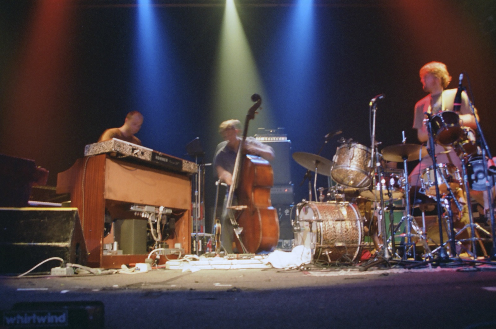 An organist, an upright bassist and a drummer play under red, blue and green light beams, seen from the stage floor.